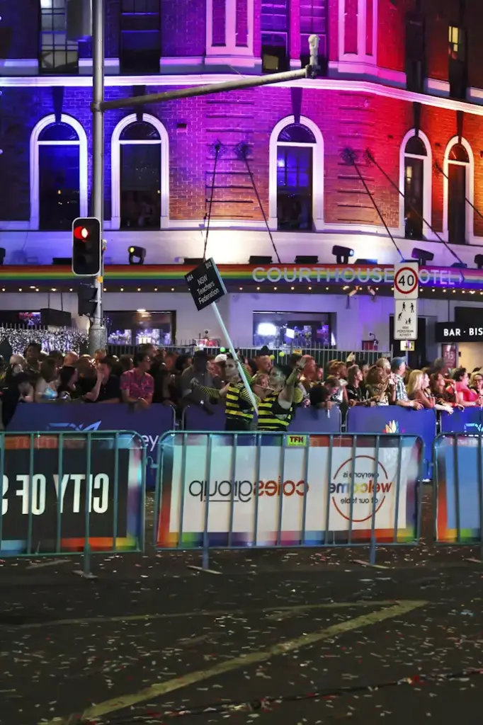 A large crowd gathers behind barricades at night outside the Courthouse Hotel, which is illuminated with vibrant purple and red lighting, while a parade participant holds a sign that reads "NSW Teachers Federation."