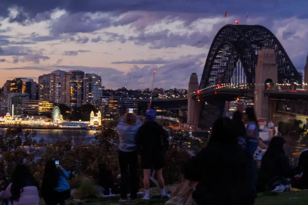 An evening view from a grassy hill showing a crowd of people looking toward the illuminated Sydney Harbour Bridge and city skyline under a twilight sky, with Luna Park's Ferris wheel visible across the water.
