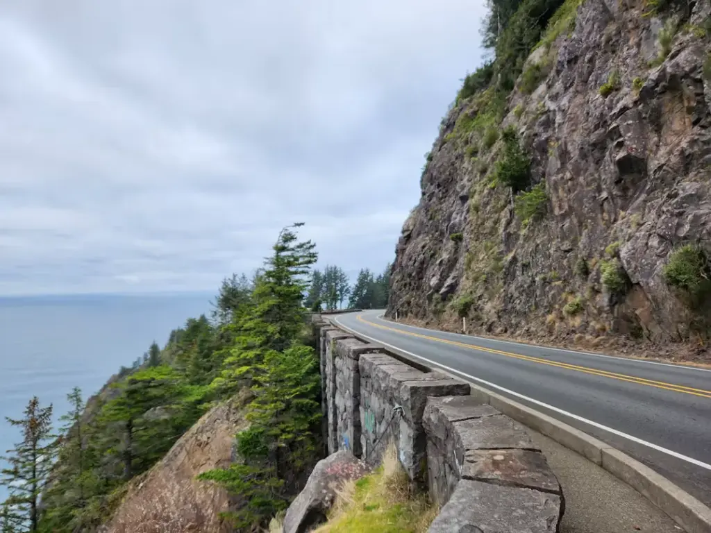 A winding coastal highway cut into a steep, rocky cliffside with a stone guardrail overlooking the ocean under a cloudy sky.