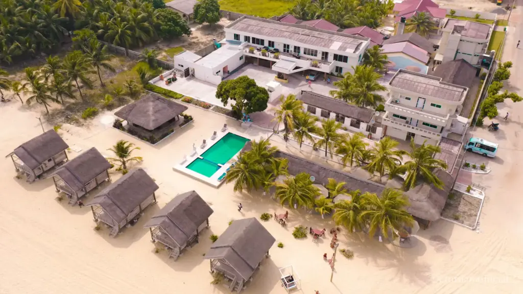 An aerial view of a beachfront resort featuring a rectangular turquoise swimming pool, rows of traditional thatched-roof huts on the sand, palm trees, and several white multi-story guest buildings.