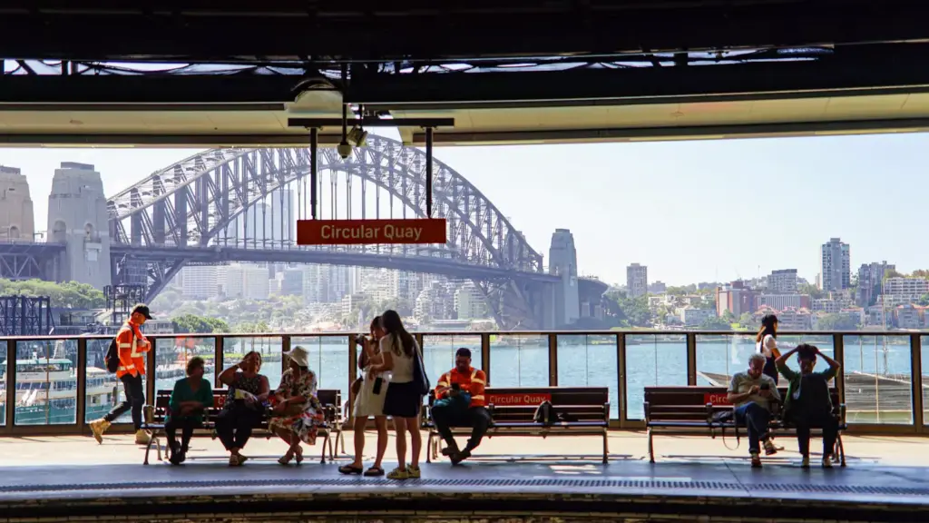 Passengers waiting on an elevated platform at Circular Quay railway station, with a hanging station sign framing a scenic background view of the Sydney Harbour Bridge over the water.